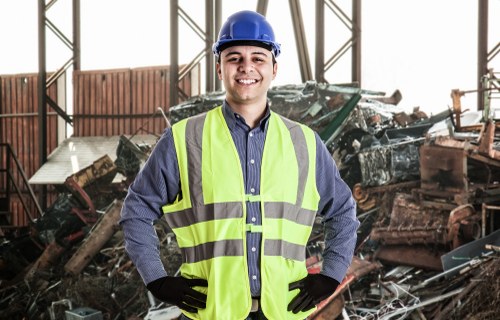 A young male waste management worker wearing a blue hard hat, a high-visibility yellow vest, a long-sleeved blue shirt, and black gloves stands in an industrial warehouse environment. Behind him, there is a large pile of mixed construction and garden waste, including wooden planks, metal debris, and plastic materials, indicating a rubbish removal site in an urban area such as Tooting. The background features a partially open metal structure with natural light illuminating the scene, highlighting the textured surfaces of the waste materials. The worker is smiling confidently, with hands on hips, suggesting a professional and approachable service provided by Garden Clearance Tooting for rubbish collection and clearance purposes, situated in the South London area, possibly within the postal district of SW17.