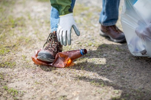 Close-up of a person wearing white gloves and brown work boots, picking up a discarded glass bottle from a patch of ground outdoor area. The bottle is transparent with a brownish tint, and is positioned near a clear plastic bag that is partially visible on the right side of the image. The ground surface consists of soil and small patches of grass, indicating a garden or outdoor space in Tooting. The background shows blurred grass and dirt, with the focus on the hand and the bottle. This scene is part of a garden clearance activity managed by Garden Clearance Tooting, highlighting the process of removing rubbish, such as glass waste, from outdoor environments.