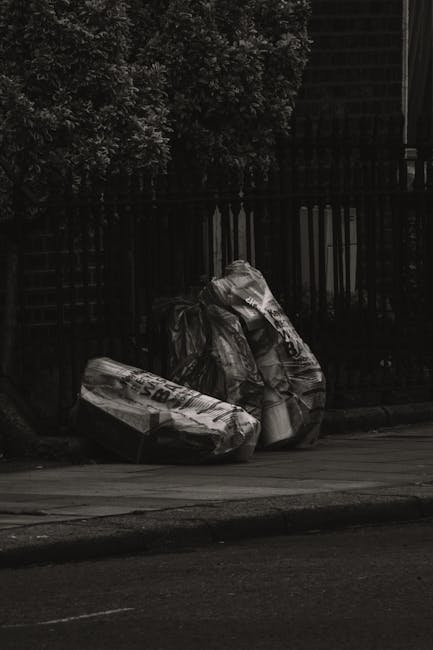 Two large black garbage bags filled with waste materials are placed on the pavement alongside a wooden fence and dense foliage. The bags appear to contain mixed refuse and are slightly crumpled, with 