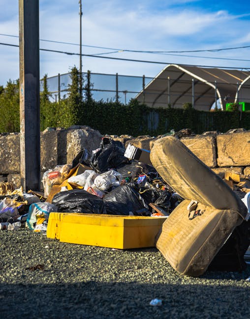 A pile of mixed rubbish, including black plastic garbage bags, loose paper and packaging, and a yellow plastic container, is scattered on gravel near a stone wall and a metal pole. An overturned, worn