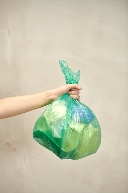 A pile of mixed rubbish, including black plastic garbage bags, loose paper and packaging, and a yellow plastic container, is scattered on gravel near a stone wall and a metal pole. An overturned, worn